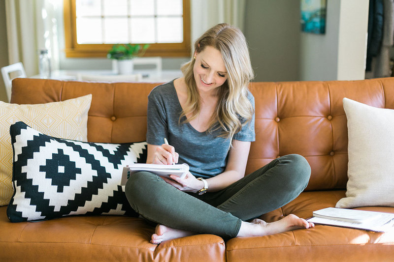woman sitting cross-legged on a tan leather couch, writing in a journal and wearing denim pants and a gray t-shirt