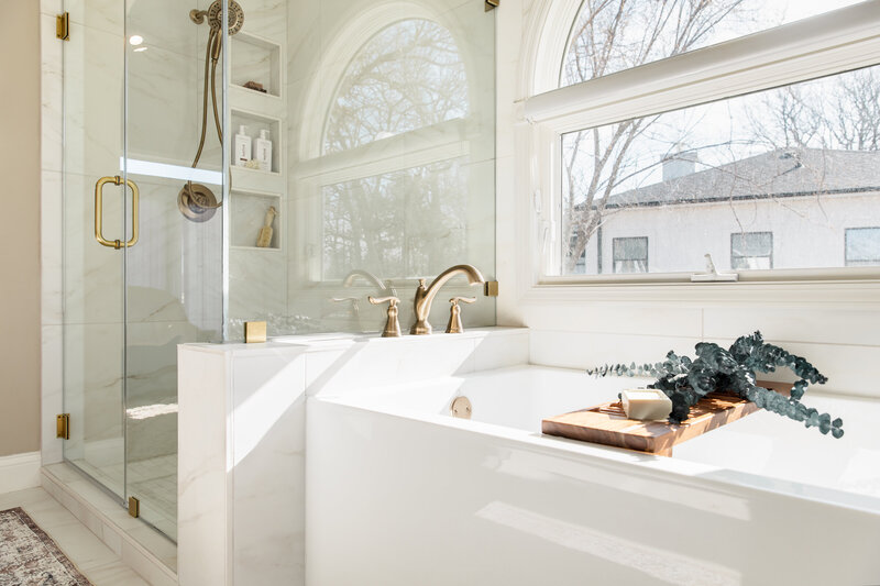Soaker tub and walk-in tile shower with large arch window. Bright and airy design with glass surround and gold fixtures.