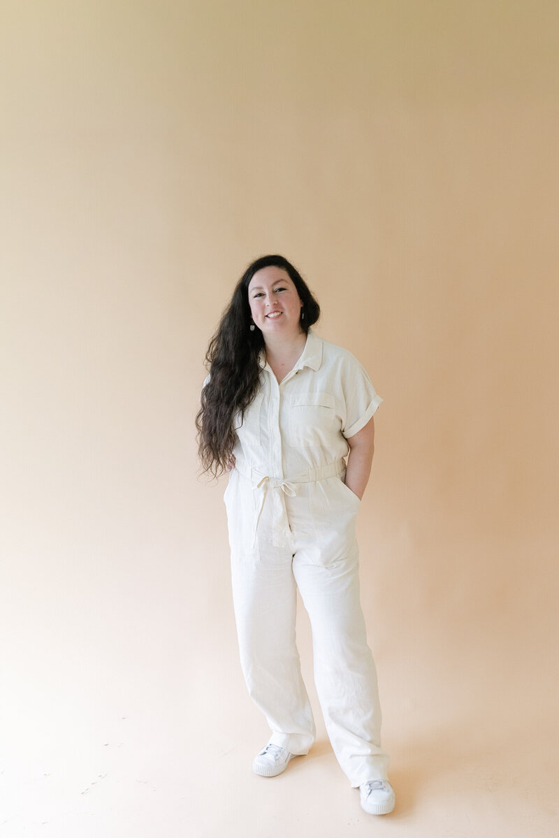 A female business owner in a white work jumpsuit smiling on a sandy colored backdrop.