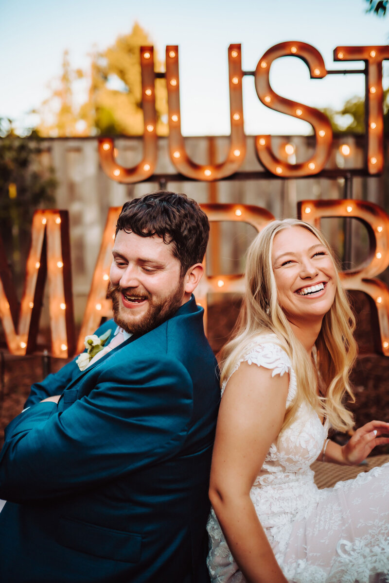 Wedding Photography, couple sitting back to back in front of "just married" sign, laughing together