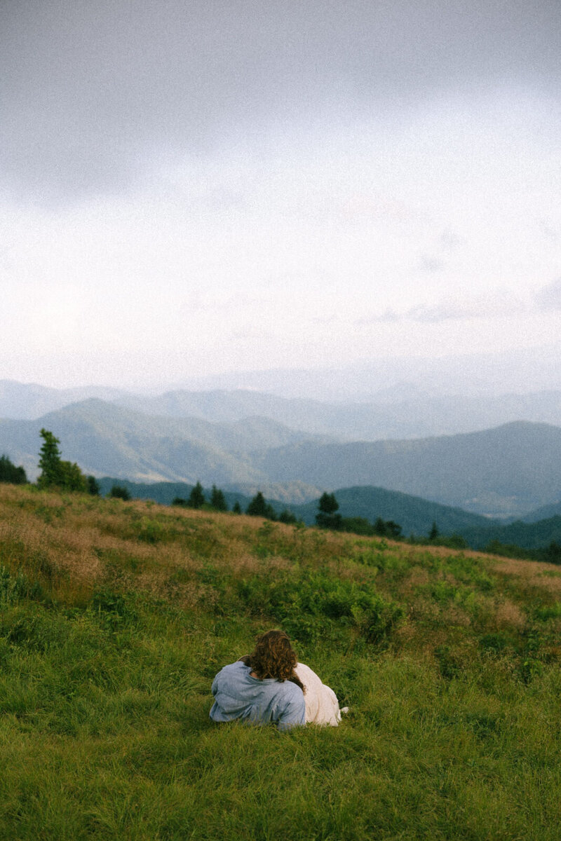A man and woman cuddling up in the Blue Ridge Mountains looking out at the view together during their engagement session in Asheville, NC with Andrea-Marie Photography