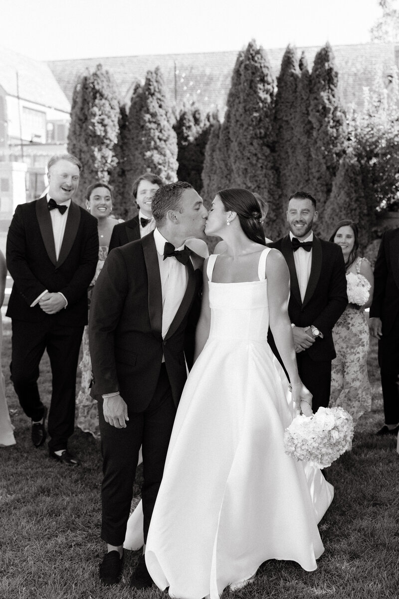 Photo of bride and groom kissing while bridal party stands behind them smiling taken by Destination wedding photographer Maine coast