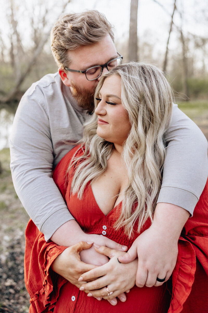 Outdoor maternity photo of parents-to-be standing close together, dad kissing mom's head and mom smiling with hands on her belly.