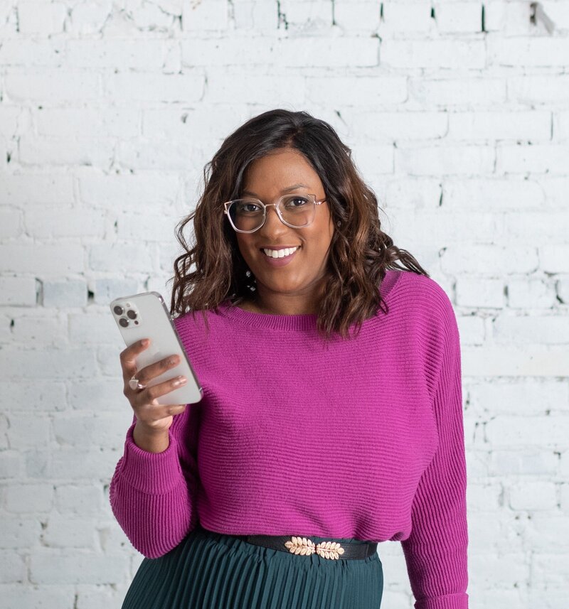 A woman with curly brown hair and glasses smiles while holding a smartphone. She is wearing a bright pink sweater and stands against a white brick wall, giving the image a fresh and modern look.