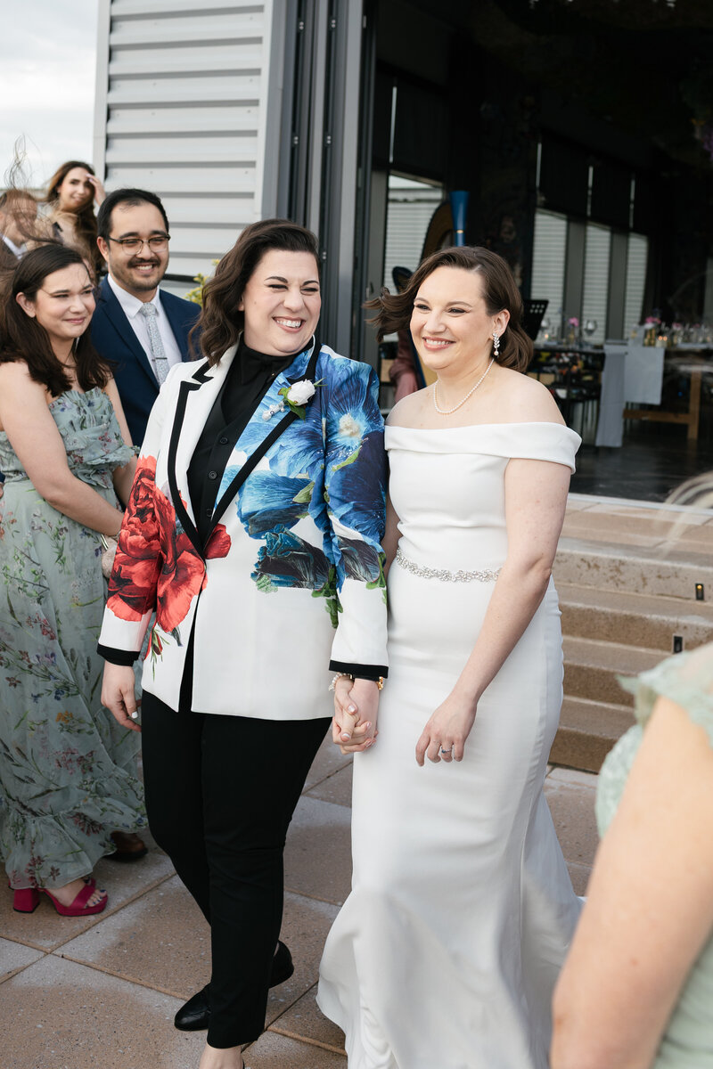 palestinian lesbian bride in a white floral tux holding hands with white lesbian bride in a white off the shoulder gown while walking to the alter together flanked by wedding guests