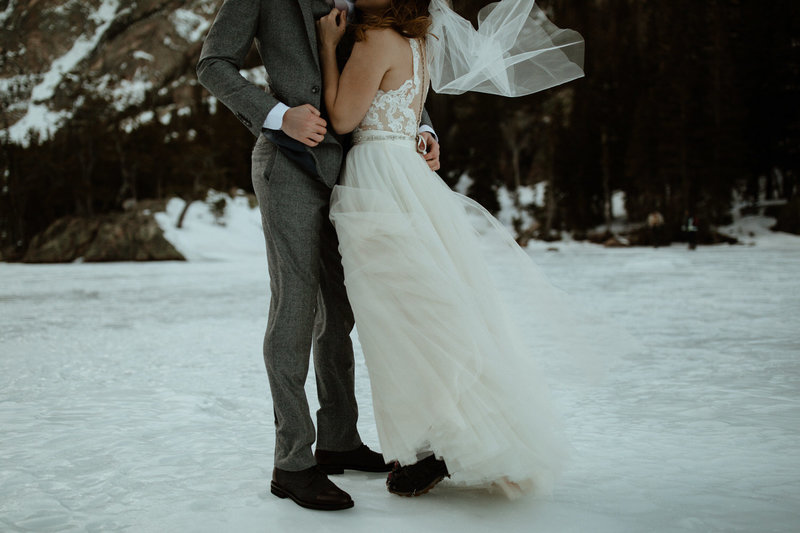 A bride and groom hold each other while a gust of wind blows against them and lifts the bride's veil, while the couple stand on snow-covered ground.