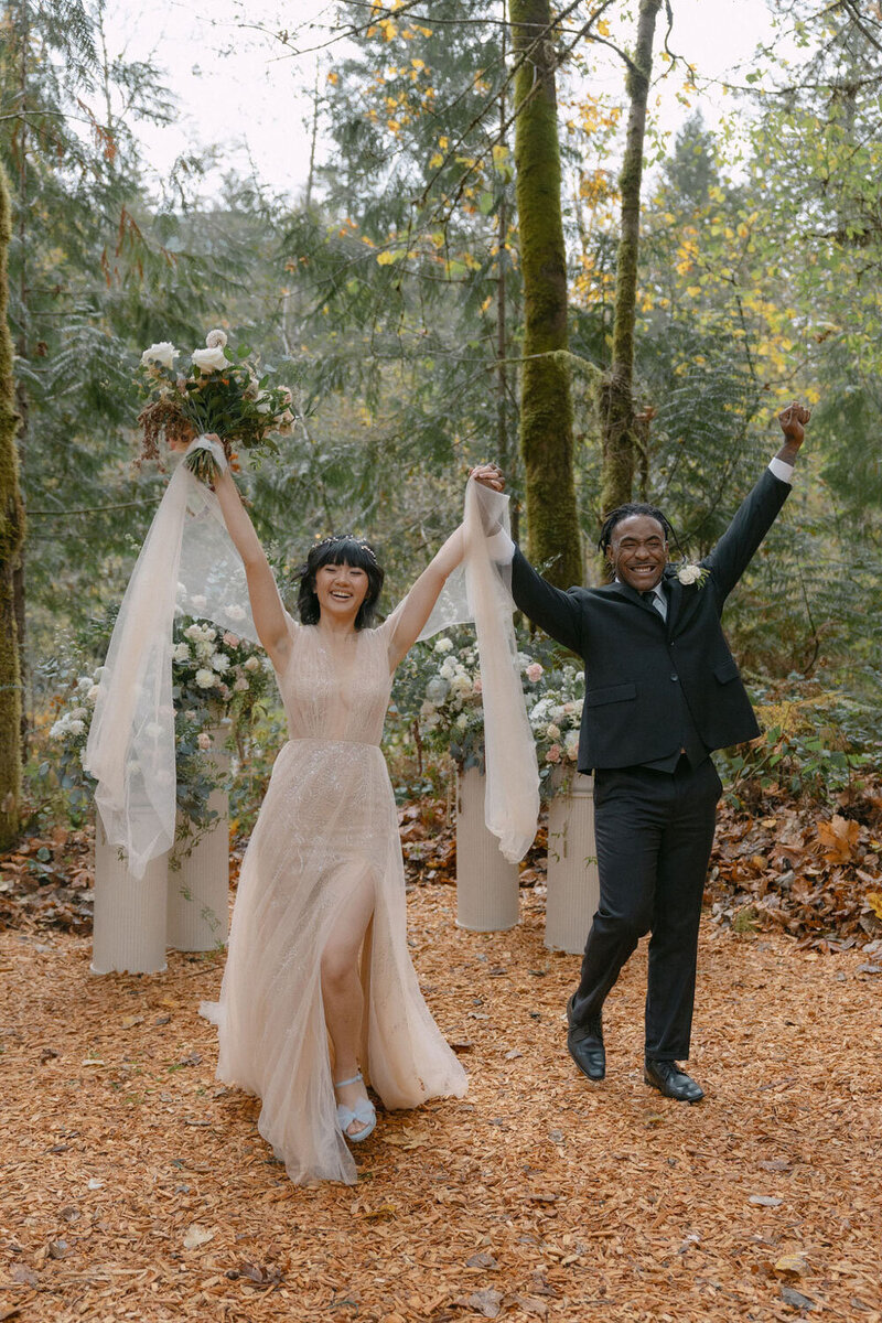 Newlywed couple celebrating joyfully with raised hands in a forest setting