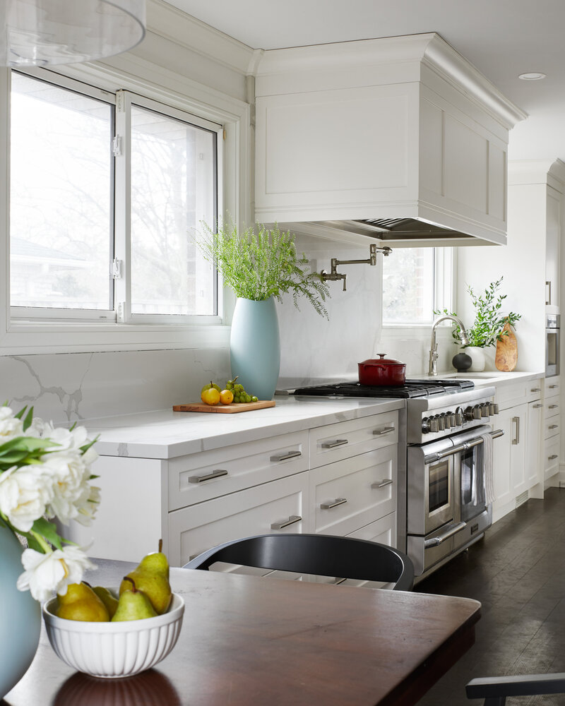 A wood kitchen table and white kitchen countertop surrounded by elegant white cabinetry and covered with small potted plants and bowls and platters of fresh fruit