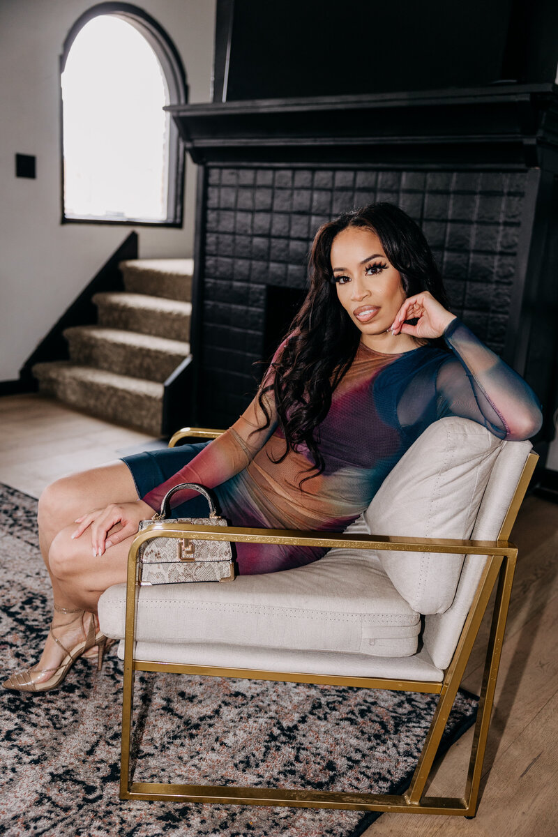 Tiara in a multicolored dress sitting with her arm on the back of a white and gold chair in front of a black fireplace