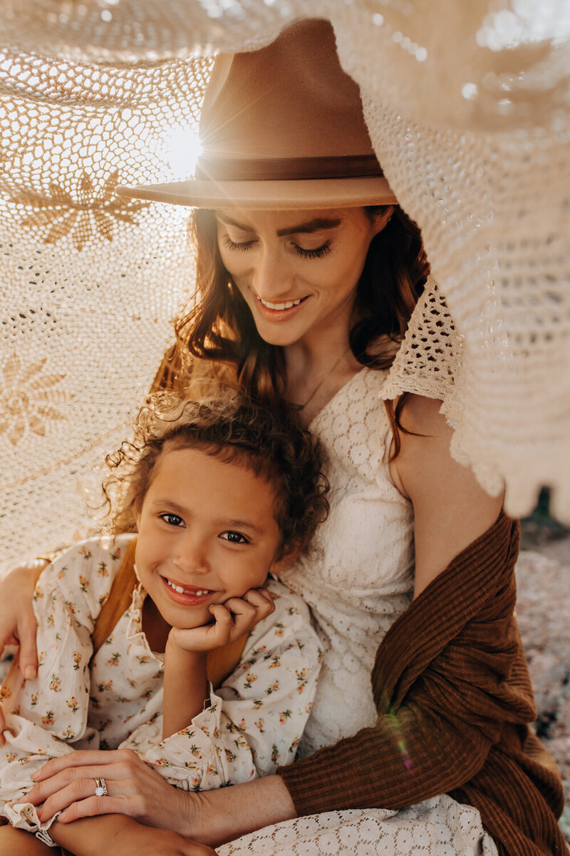 A mom smiling down at her daughter under a knit blanket. The sun behind them barely peeks through.