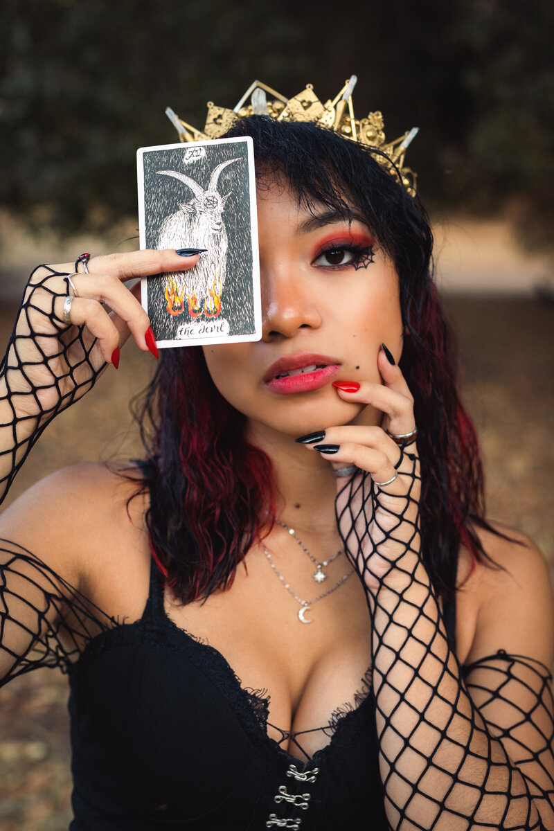 person in a gold crown holding a tarot card up over one eye