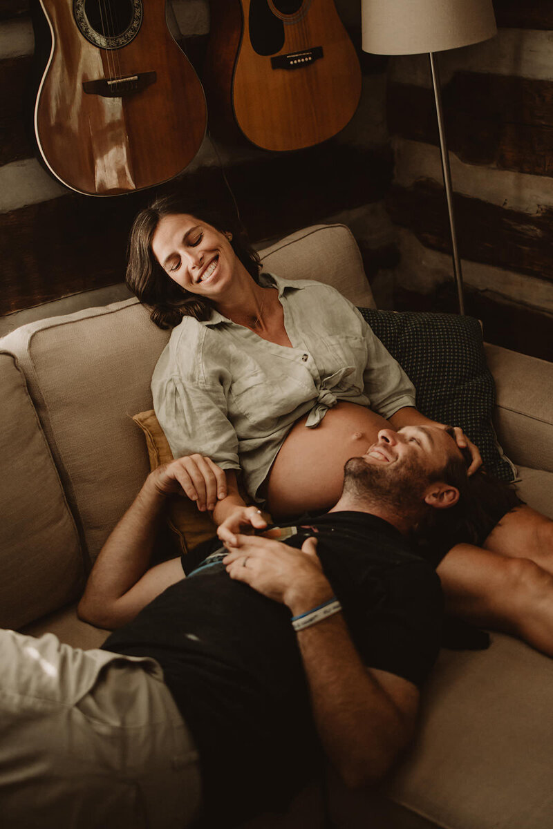 A pregnant person sitting on a couch with their partner laying on their lap as they smile at each other 