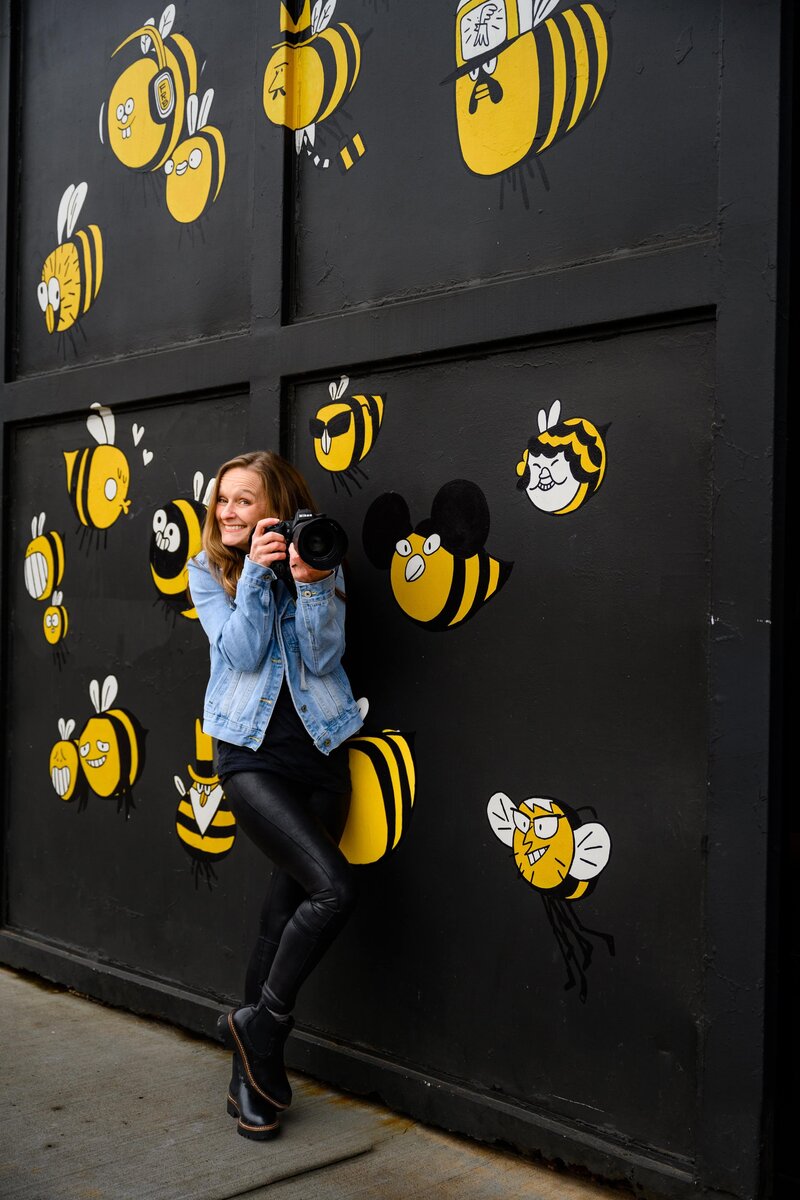 Charlotte, NC photographer in a denim jacket and black pants holding a camera stands against a wall decorated with various cartoon bees.