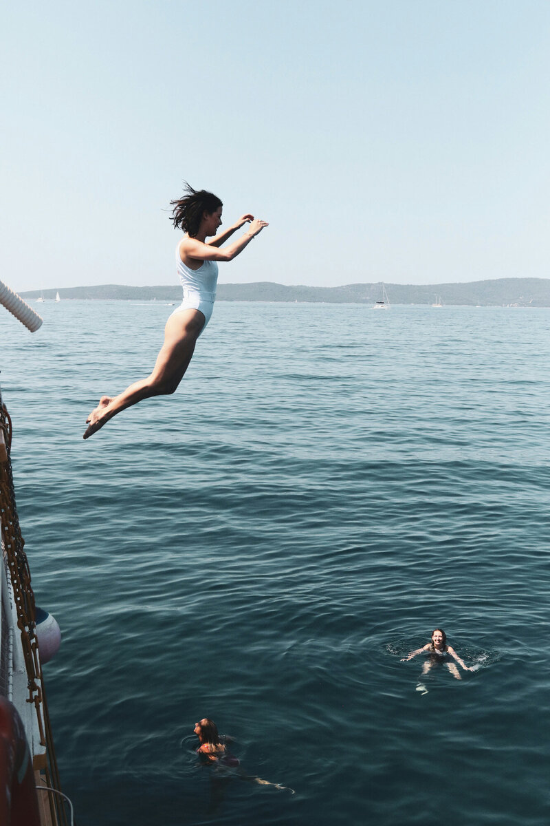 person in a white swimsuit is captured mid-air, jumping off a boat into the sea. Two other swimmers are visible in the water below