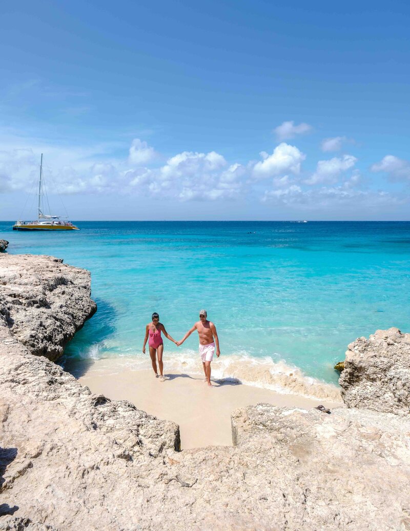 A couple holding hands walks from the turquoise water onto a small sandy cove surrounded by rocky cliffs, with a sailboat anchored in the distance under a bright blue sky.