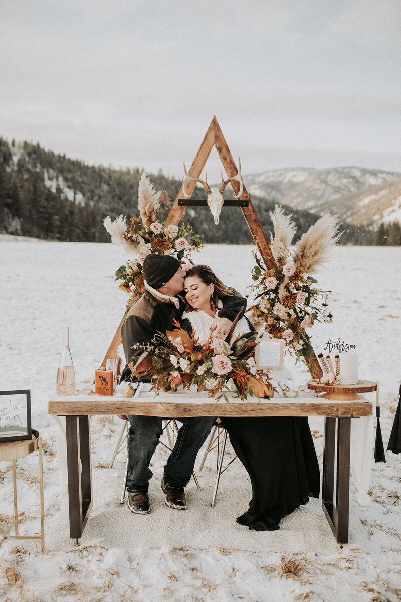 A man kisses his boho bride while sitting at a table in the mountains