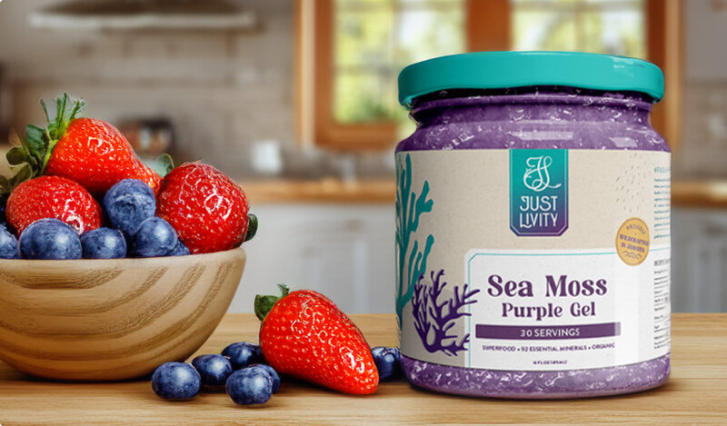 Mason jar with purple gel with packaging label for the brand Just Livity. The label showcases an underwater sea theme of sea moss. On the left side of the jar is a bowl of strawberries and blueberries. Both these items sit on a kitchen counter.