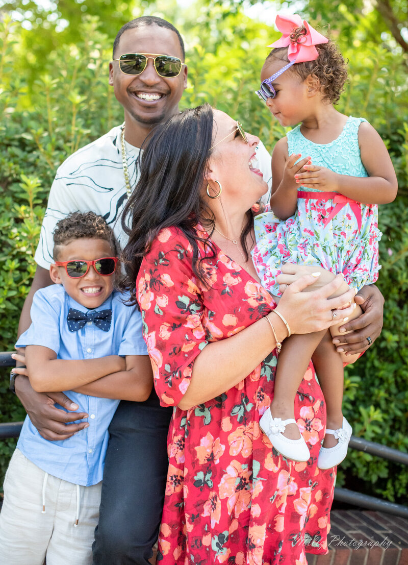 a family of four, all wearing sunglasses. the father has his arms wrapped around their son while the mother is holding their daughter laughing and smiling at her. photographed by Millz Photography in Greenville, SC