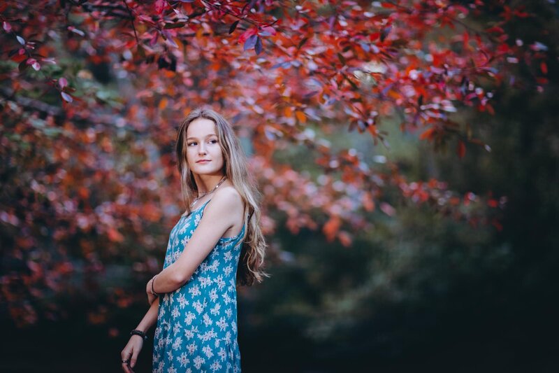 Moody senior portrait of a girl standing in front of gorgeous red fall leaves looking over her shoulder.