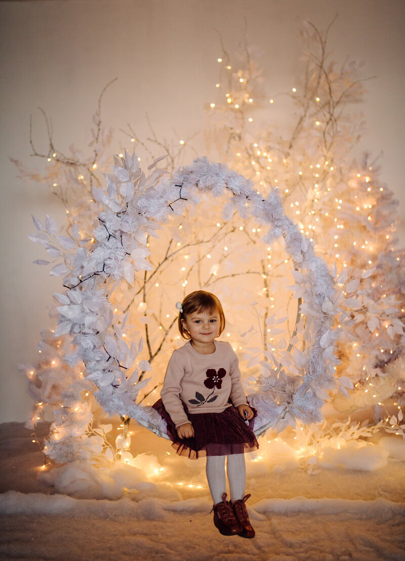 Petite fille assise sur une arche ronde flottante décorée de feuilles enneigées. Décor féérique crée par une event planner.