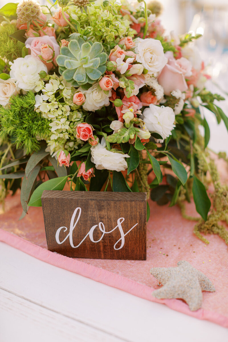Lush floral centerpiece with roses and succulents on wooden block with 'dos' text.