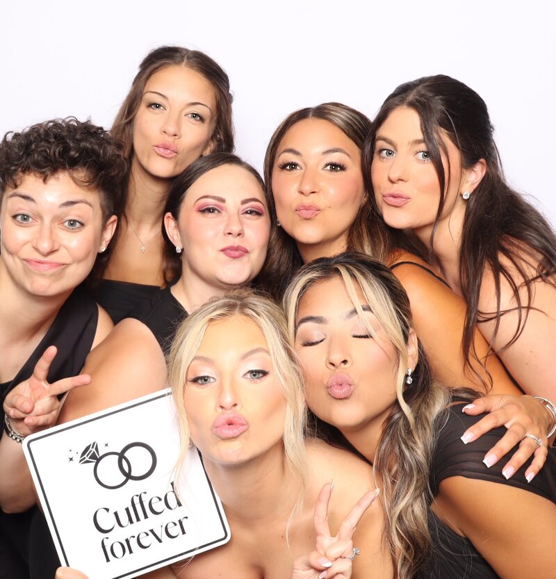 Three women standing together and laughing in front of a white brick wall at a natural light photo booth, dressed casually for a fun and relaxed event.