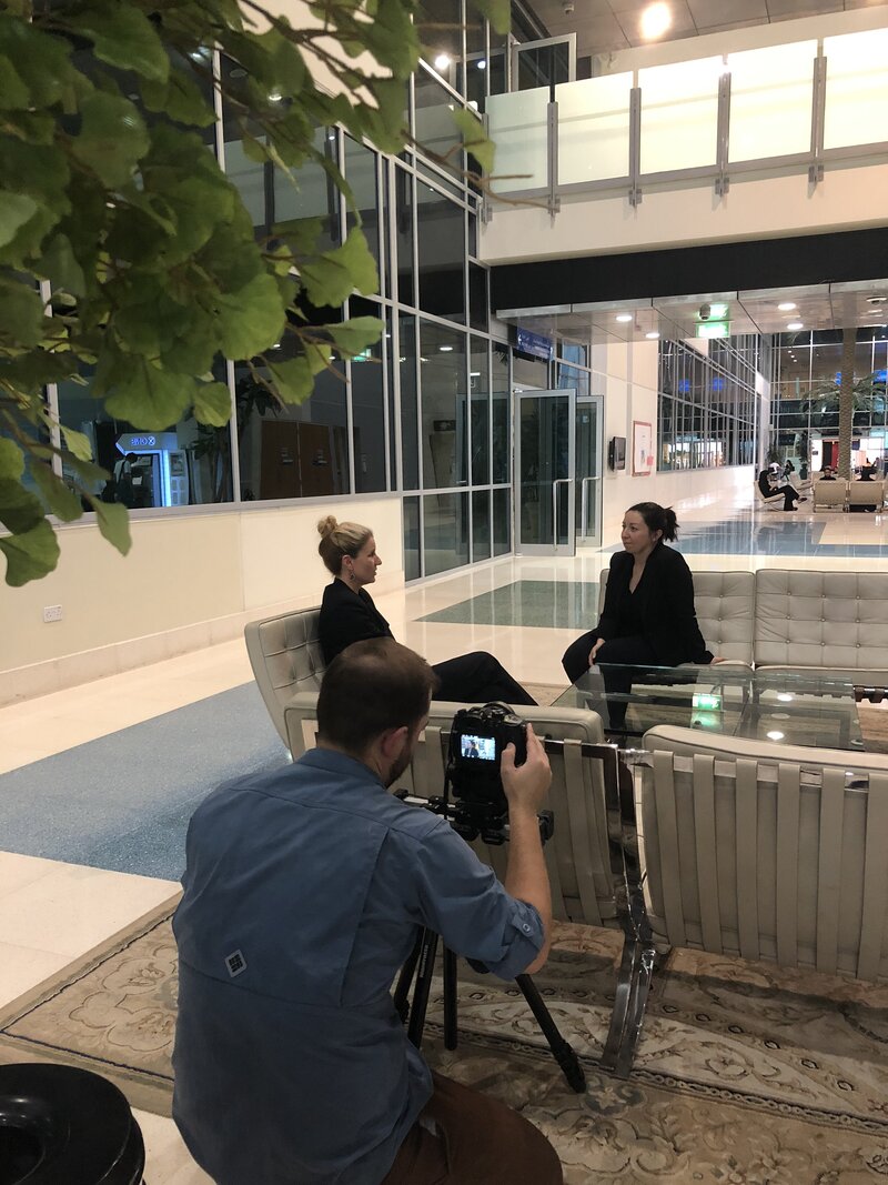 Two people are having an interview in an airy, glass-walled indoor space while a cameraman records the conversation, focusing on educational leadership and the impact of coaching.
