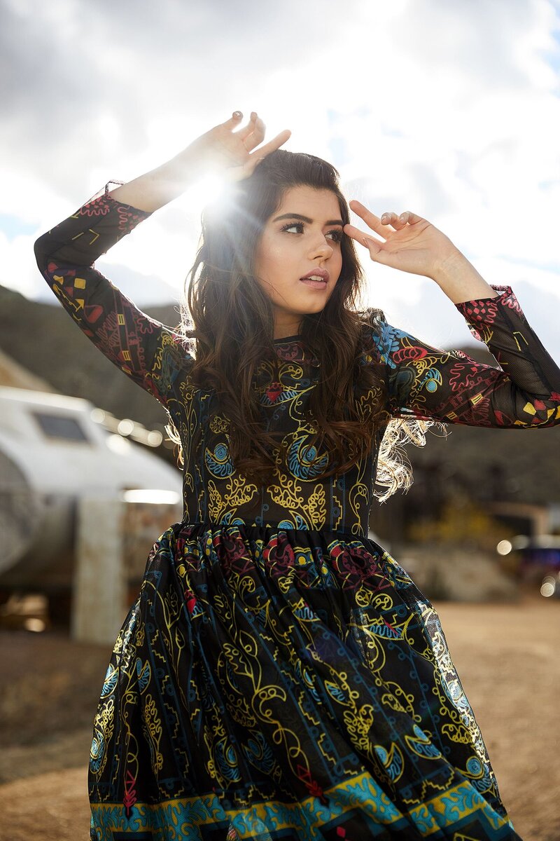  Editorial-style teen model wearing a sheer, patterned black dress, posing outdoors with the sun beaming behind her during a high-fashion styled shoot with Tara Rochelle.