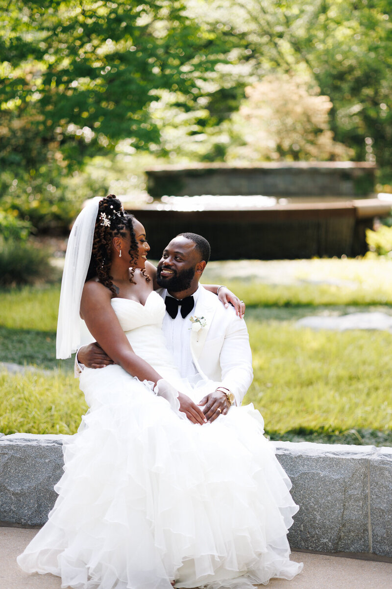 A bride sitting on the lap of a groom inside of Atlanta botanical garden looking at each other smiling.