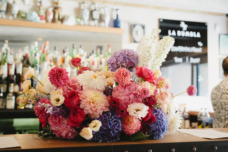 Colorful floral arrangement with roses, dahlias, and hydrangeas in shades of pink, purple, red, and white, displayed on a bar counter with bottles and glassware in the background. By Melissa Clark Photography