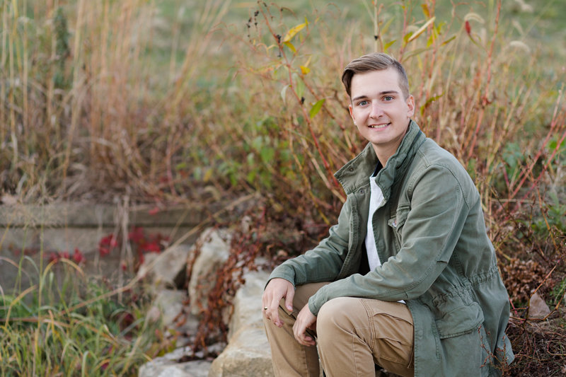 perry high school senior guy sitting among tall grass, phperry high school senior guy sitting on concrete ledge, among tall grasses in fall, so lots of color, hes looking at camera, photographed by Jamie Lynette Photography canton ohio senior photographer
