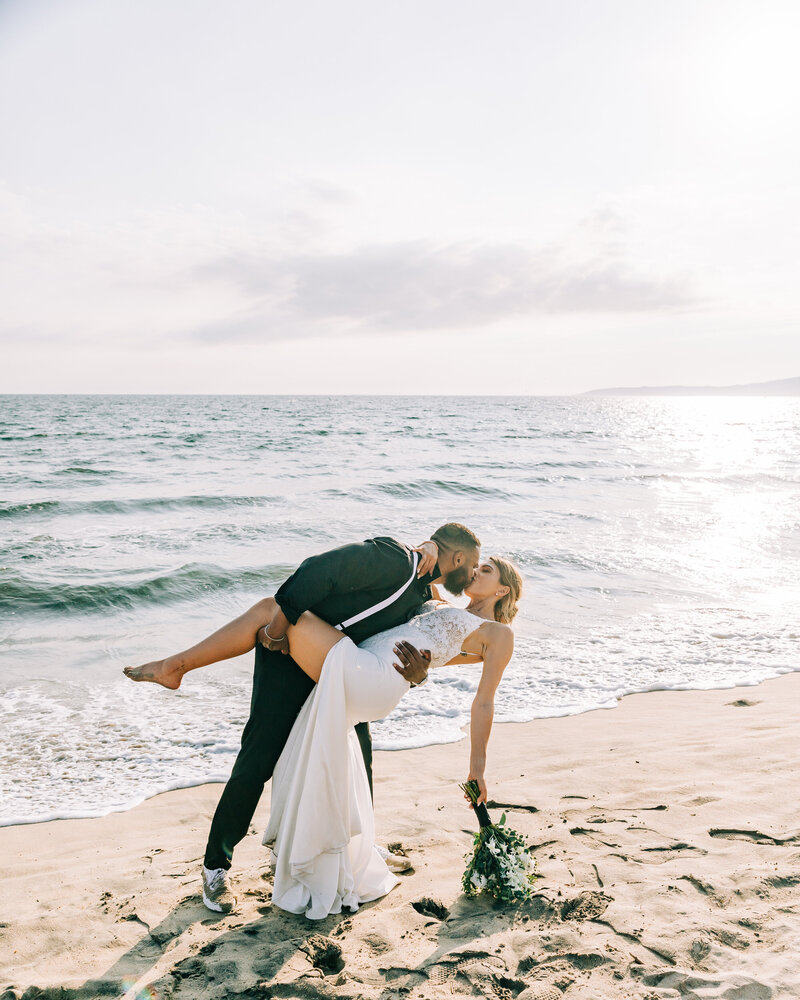 A groom lifts his bride as they share a kiss on the beach. The bride wears a white gown with a slit, and the groom is dressed in dark attire. The ocean and sky create a serene backdrop to the moment.