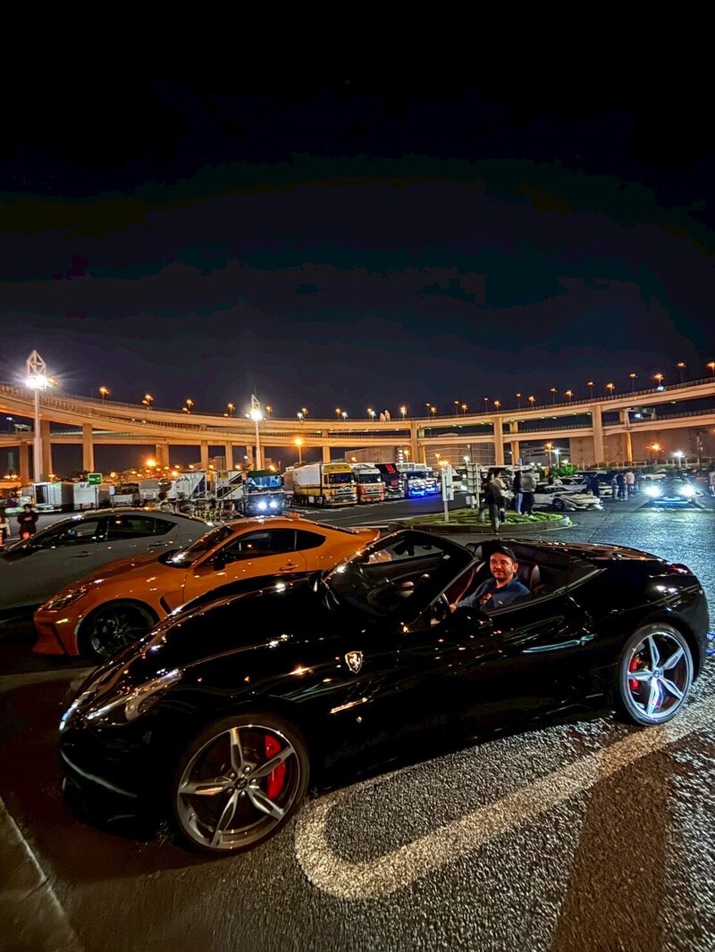 A guest of Tokyo Luxe Drive smiles inside a black Ferrari during a nighttime tour stop at Daikoku Parking Area, with luxury cars and highway loops in the background.