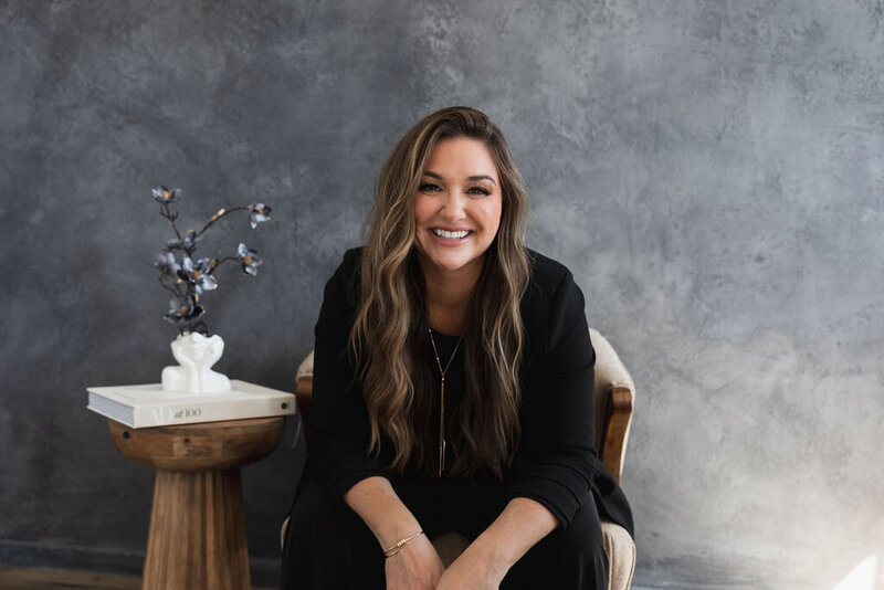 A hairstylist wearing long extensions is wearing a plazer and jeans, leaning against a wall, smiling at the camera.