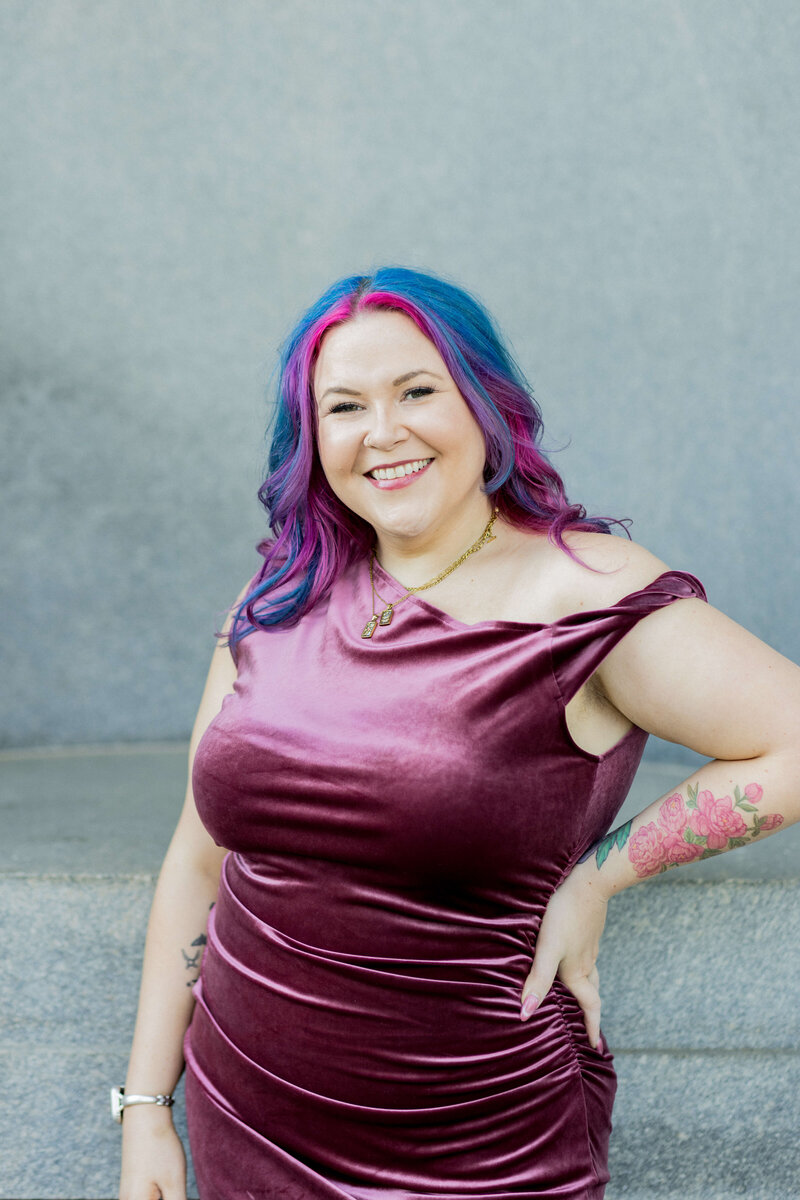 Jessica Hunt, the lead creative behind Jessica Hunt Photography, poses confidently in front of a smooth gray stone backdrop. She is wearing a rich mauve velvet off-the-shoulder dress, with her hand on her hip and a bright smile. Her vibrant hair is dyed in bold shades of pink, purple, and blue, and she wears gold jewelry and a silver watch. A detailed floral tattoo adorns her forearm, adding a personal touch to her expressive, polished look.
