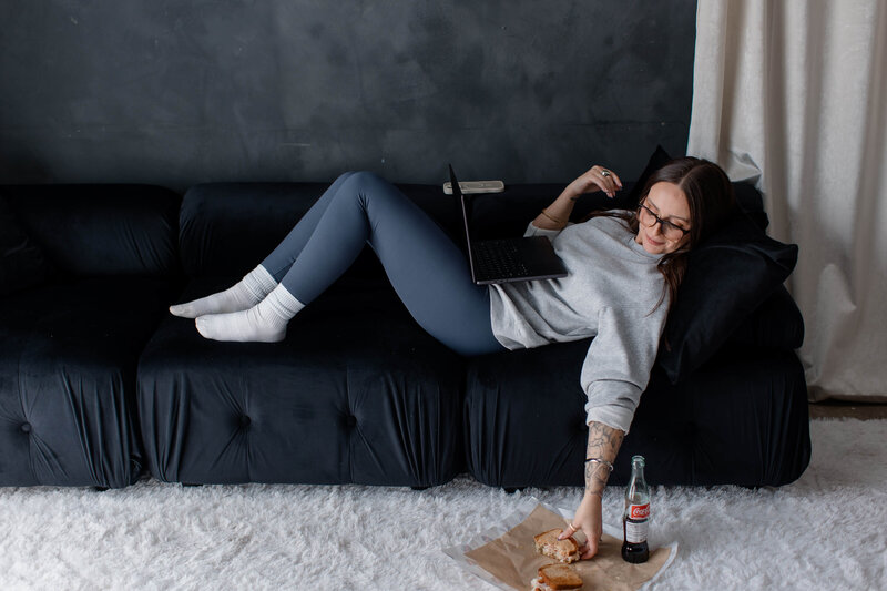 Sharon is laying on a sofa with her laptop in her lap and she's taking a moment to grab her sandwich to eat while she works.