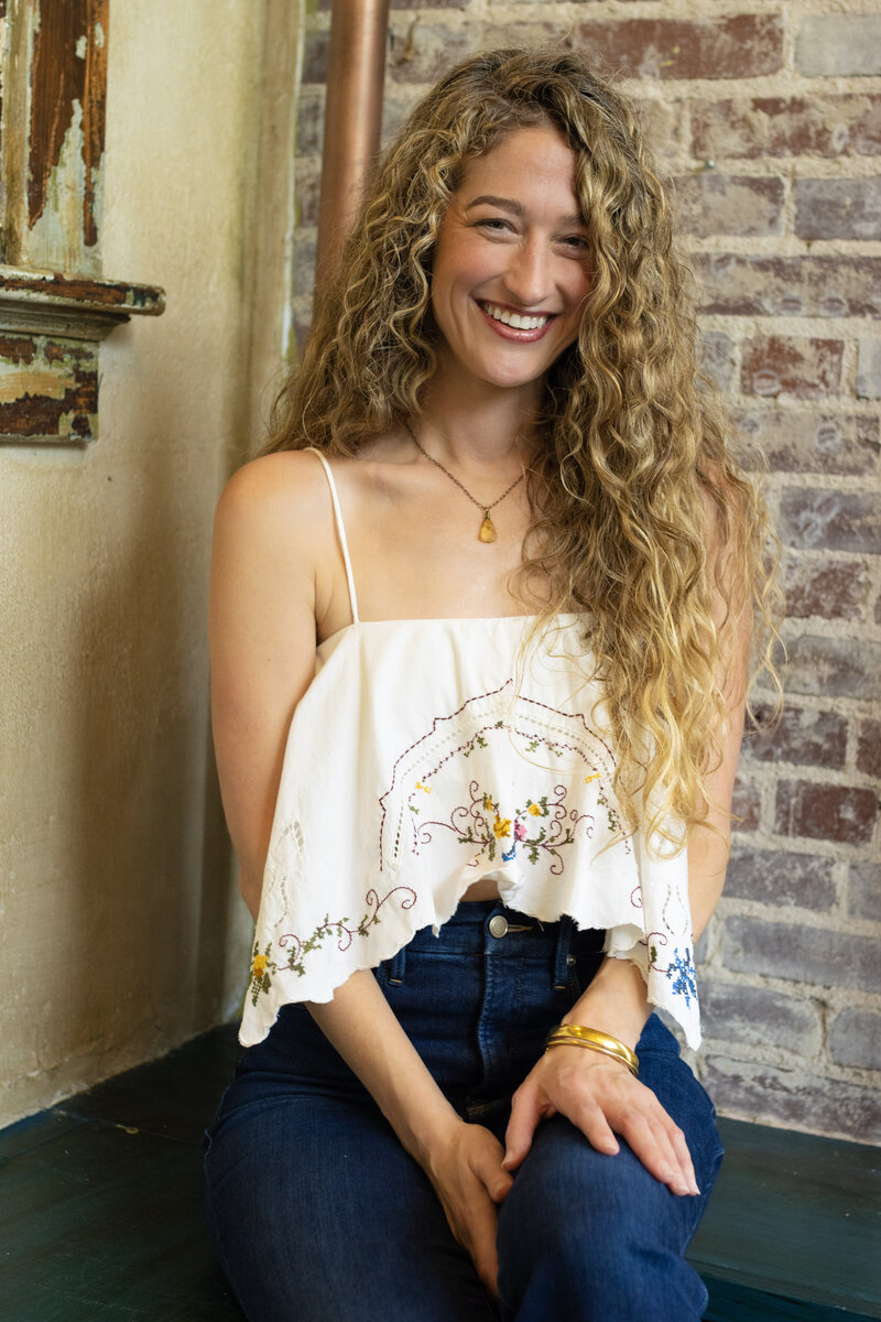 Dr. Mamie Burruss, ND, LAc smiling and sitting casually in front of exposed brick