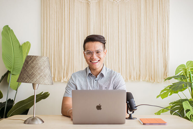 Sho Dewan, young Asian man, career coach and founder of Workhap, sitting at a desk behind his laptop, with his microphone and tablet on the table, there are large green plants on each side of him, he's wearing his glassed and looking straight at the camera with a large smile