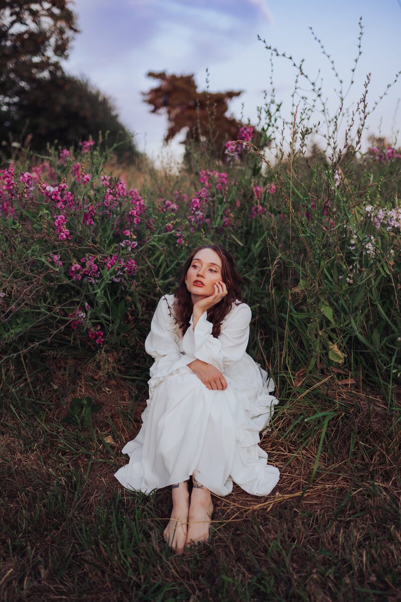 Young, red-headed woman sits in a flowy white dress in a field of flowers while looking away from the camera and posing with her head rested on her hand.