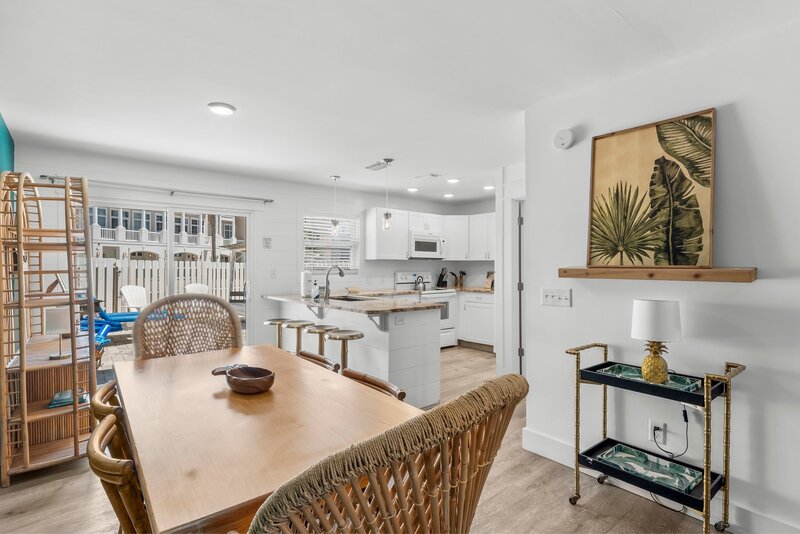Chic open-plan kitchen and dining area with white cabinetry, island seating, rattan decor, and sliding doors to private patio in PCB rental.