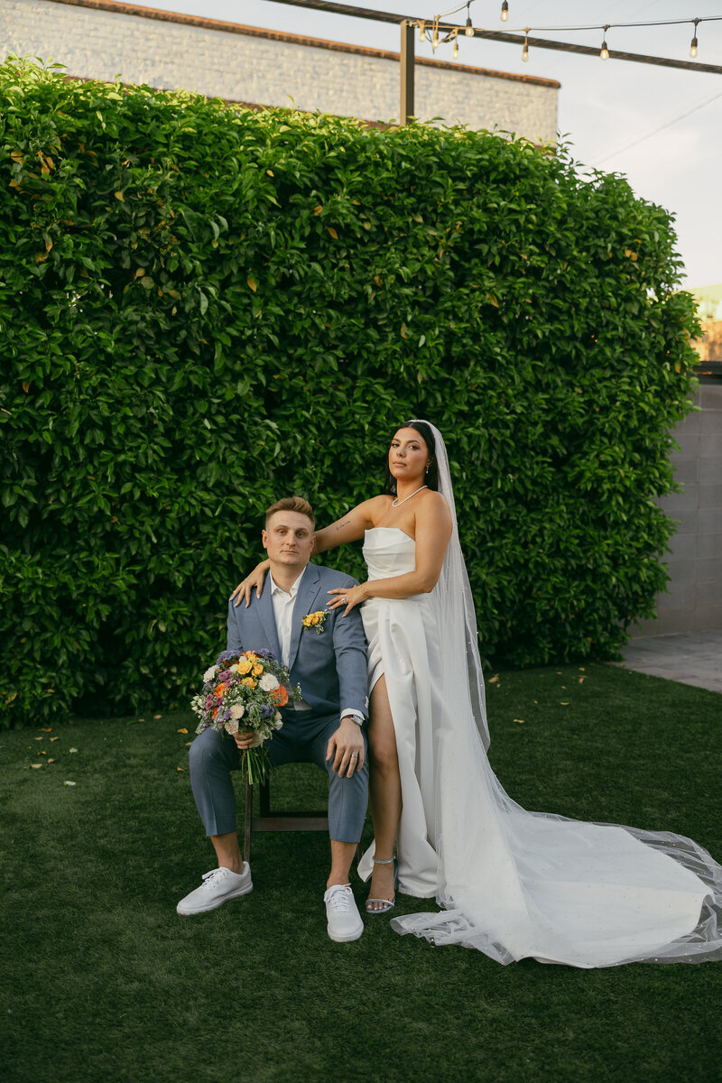 A groom sits in a chair holding his brides bouquet while the bride stands to his side with her hands draped over his shoulders while both look directly into the camera.