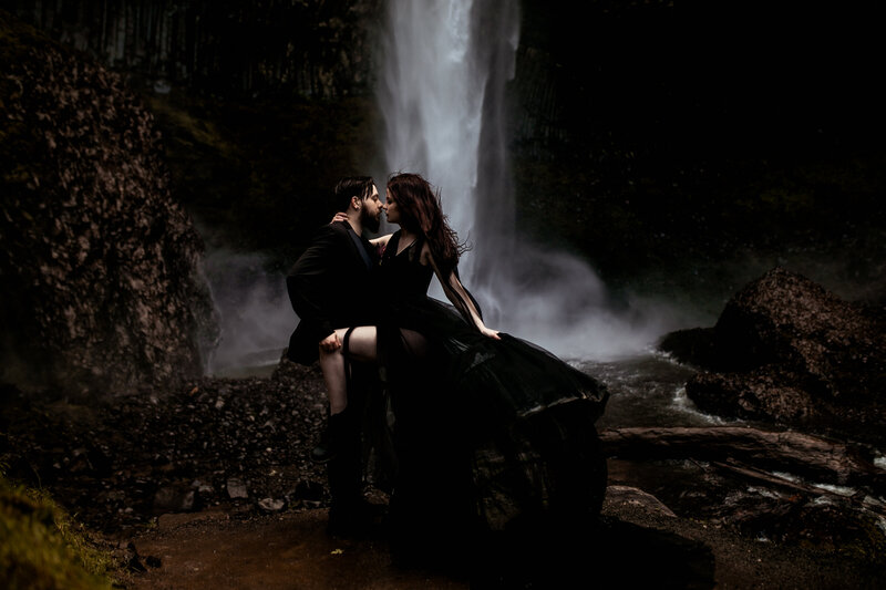 A couple dressed in black sharing a moody, romantic moment by a waterfall during their Oregon Coast elopement.