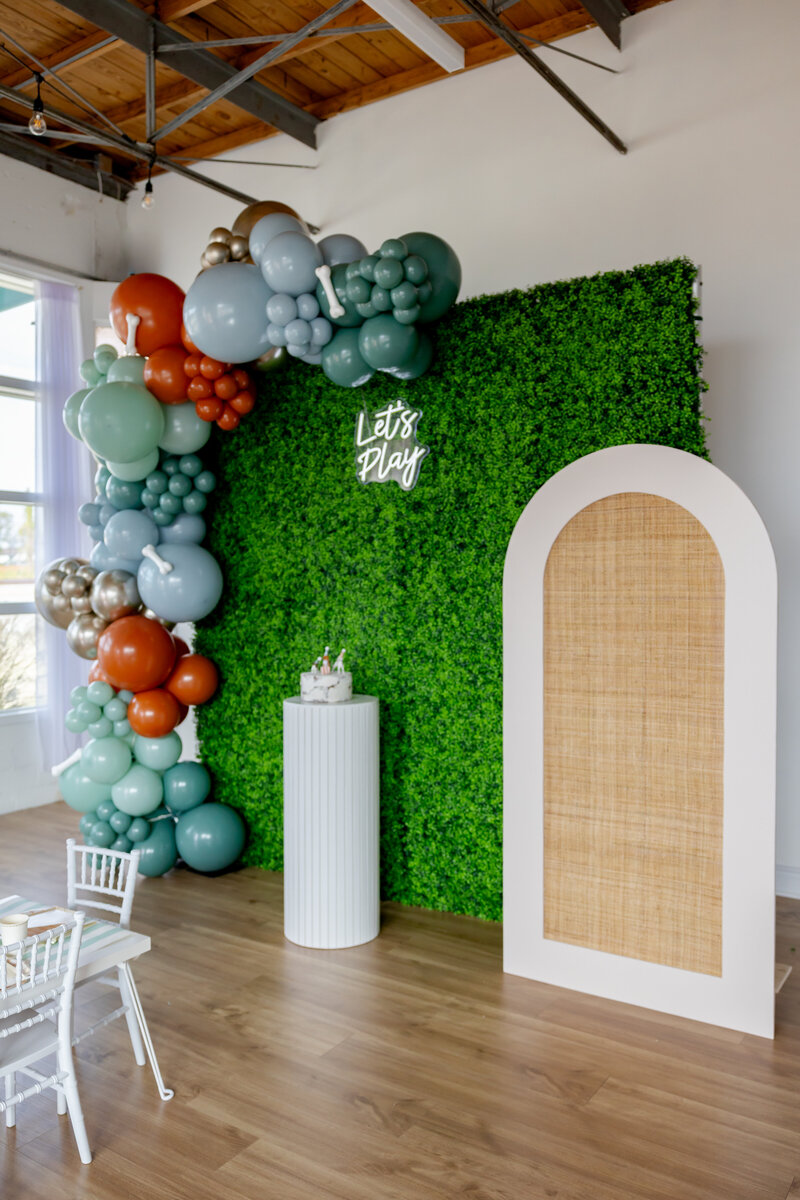 Dog party wall with rattan arch, boxwood floral wall, dog balloons, cake pedestal and dog cake.