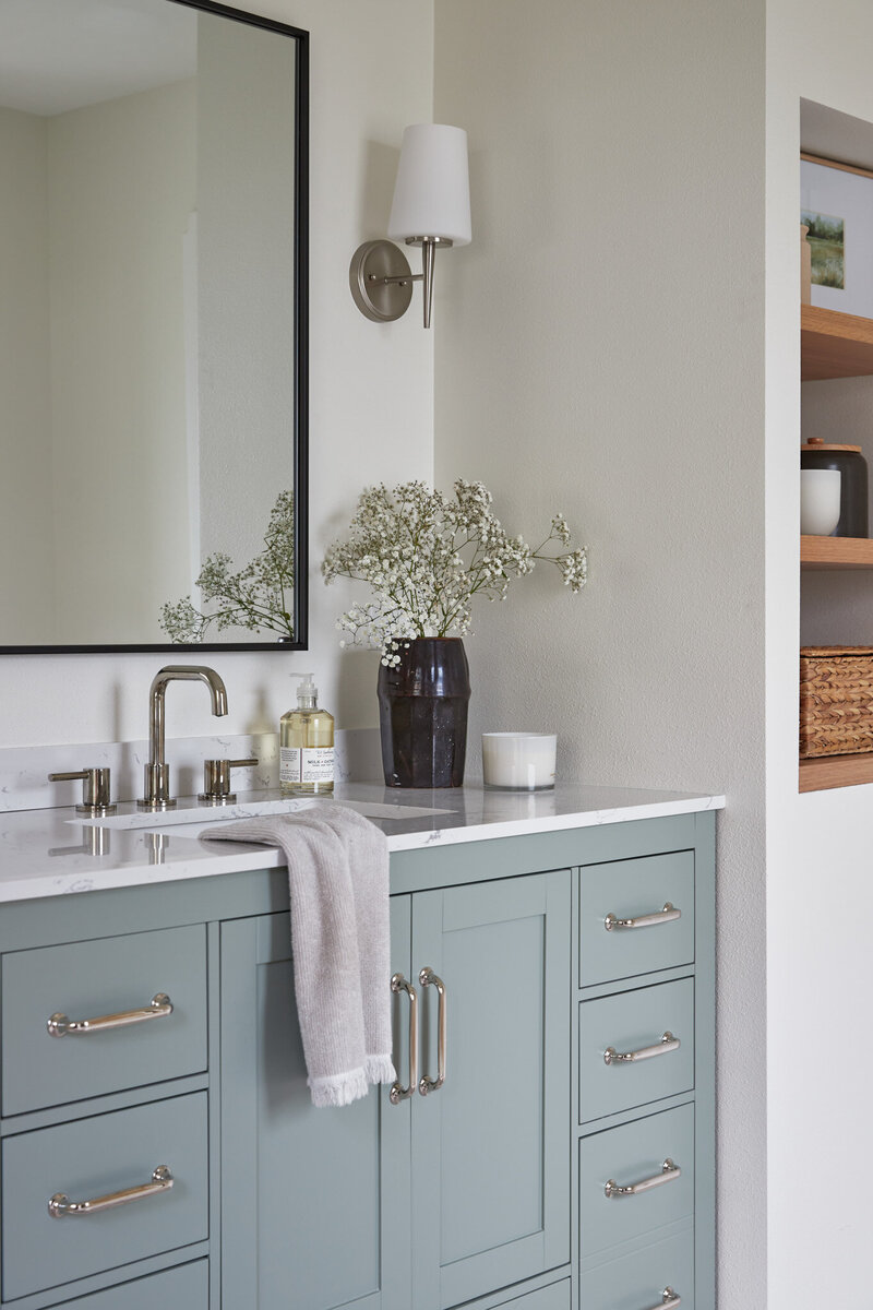 Bathroom with light blue bathroom vanity with silver cabinet pulls. A silver sink faucet with a black mirror above. Next to the sink is a dark brown vase with baby’s breath flowers, a Handsoap pump, and a white candle. Next to the vanity is a white wall with recessed white oak shelves with photos, jars, and a woven basket.