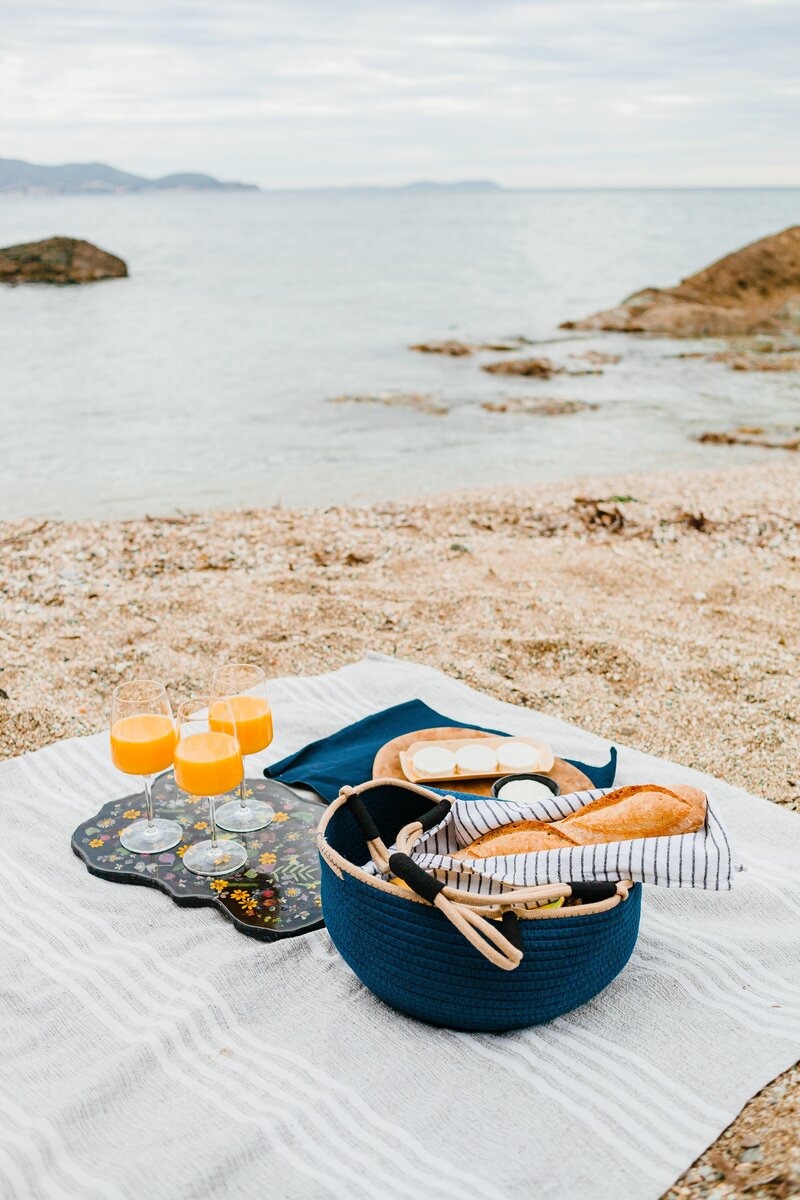 Image shows wine glasses with an orange beverage and a basket of bread on a blanket on the sand, water in the background