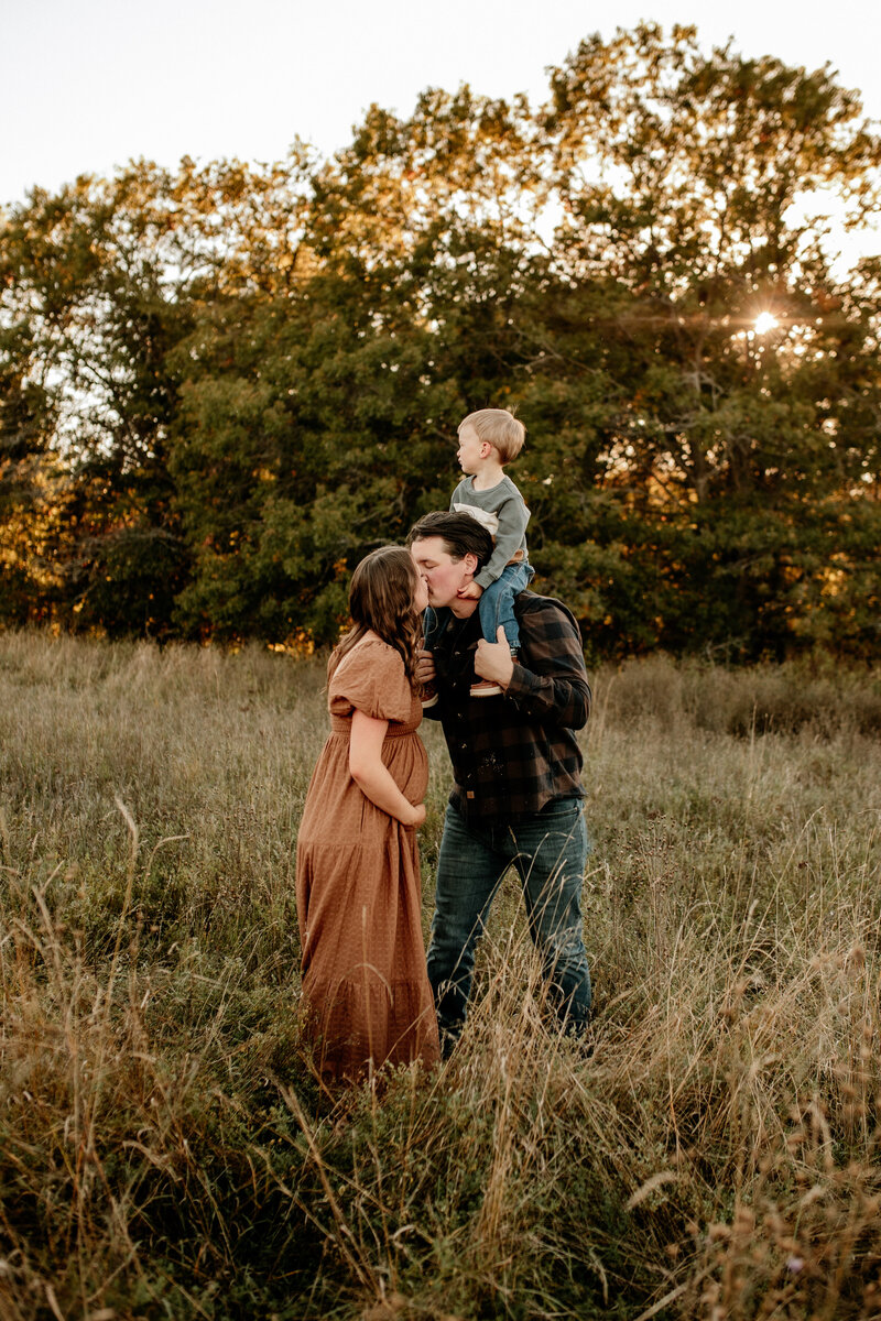 Mom and dad share a smooch while their toddler perches on dad's shoulders during a Fall pregnancy announcement photoshoot.