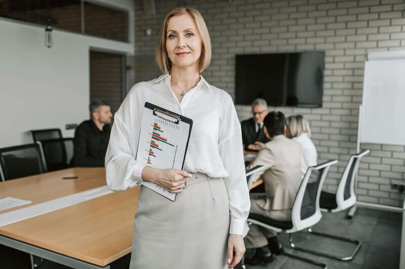 A confident woman standing in a boardroom, holding a clipboard with bar charts, while a team sits at a table in the background.