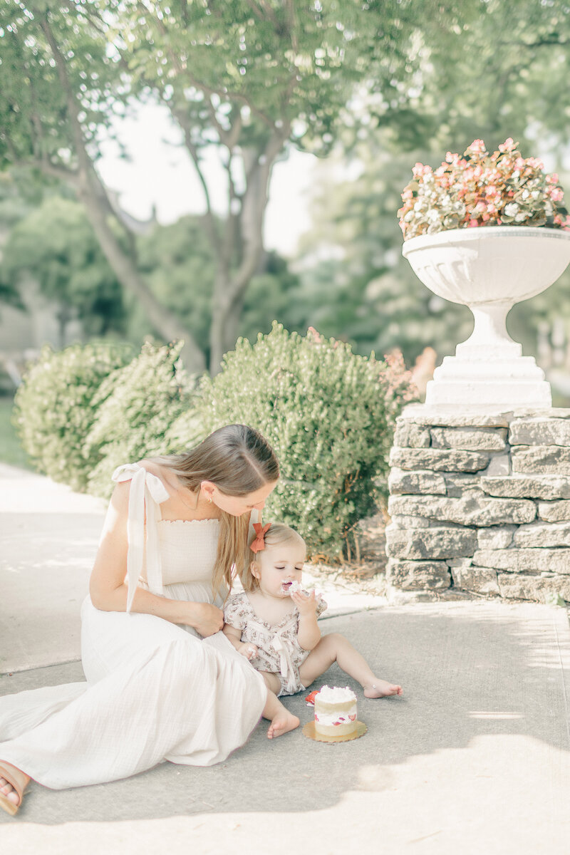 Photo of a mother sitting on the sidewalk with her one-year-old daughter enjoying a cake smash celebration in Kansas City