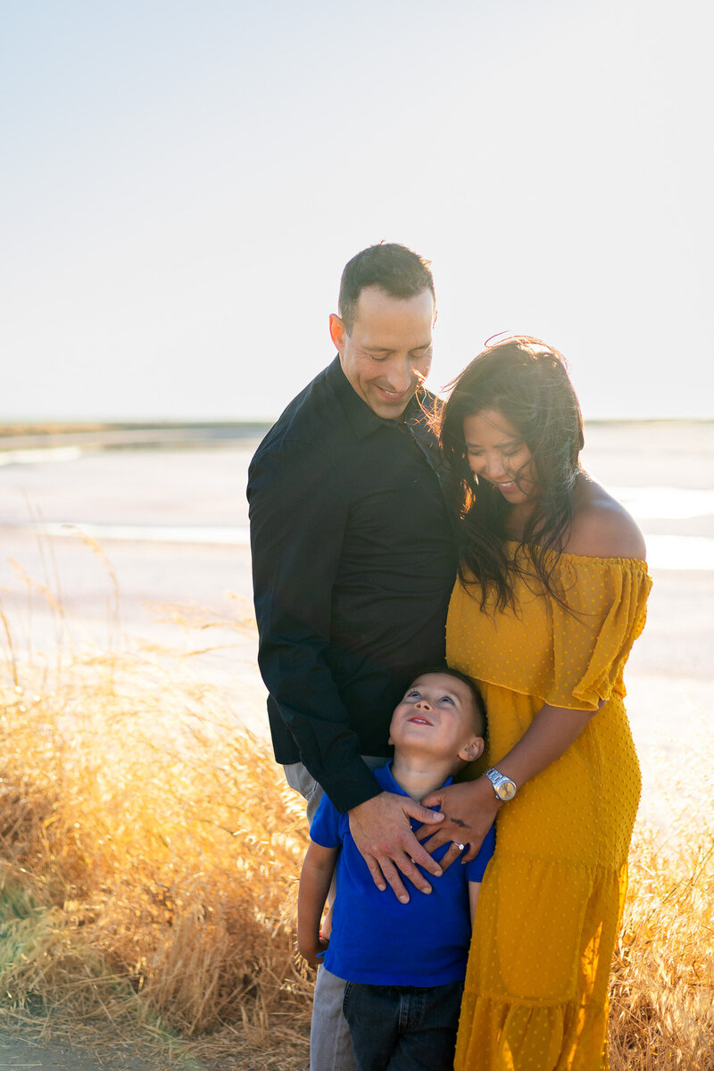 A tender family moment during golden hour with parents smiling down at their young son – Bay Area Photographer Session Info and Pricing - Ellobelle Photography