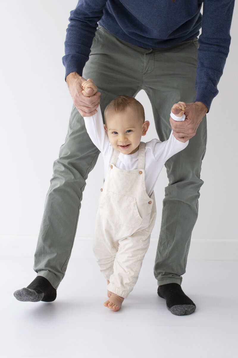 A baby in cream overalls and a white long-sleeved shirt is taking their first steps while holding an adult's hands for support. The baby smiles happily, and the adult, wearing grey trousers and dark socks, is standing behind on a white studio background.